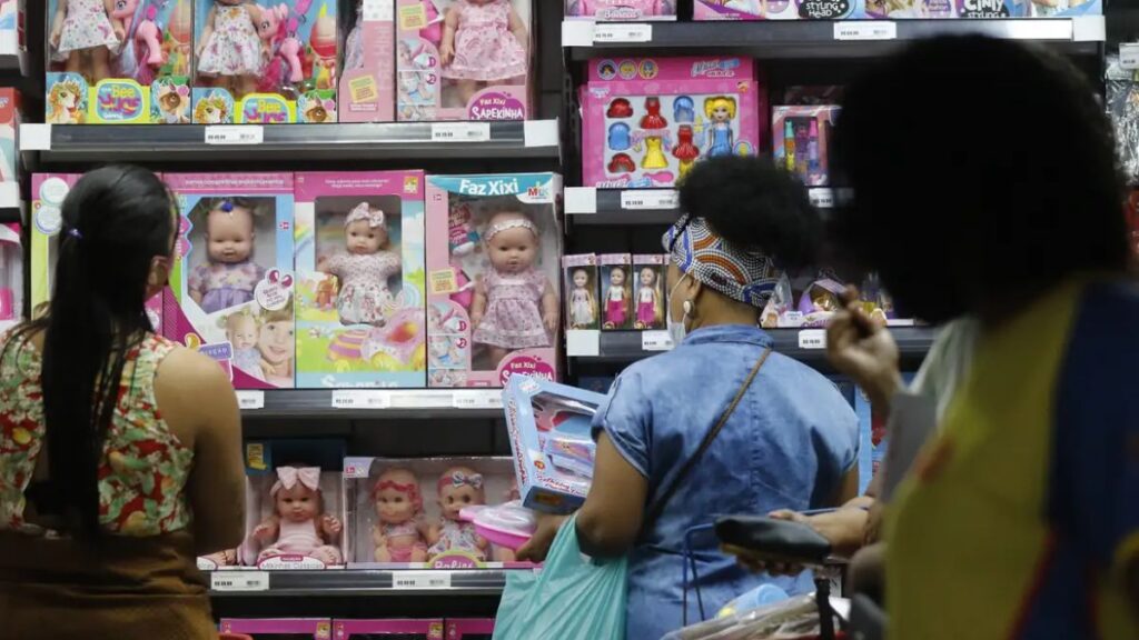 Na imagem, duas mulheres aparecem de costas, olhando a prateleira de uma loja cheia de bonecas 
