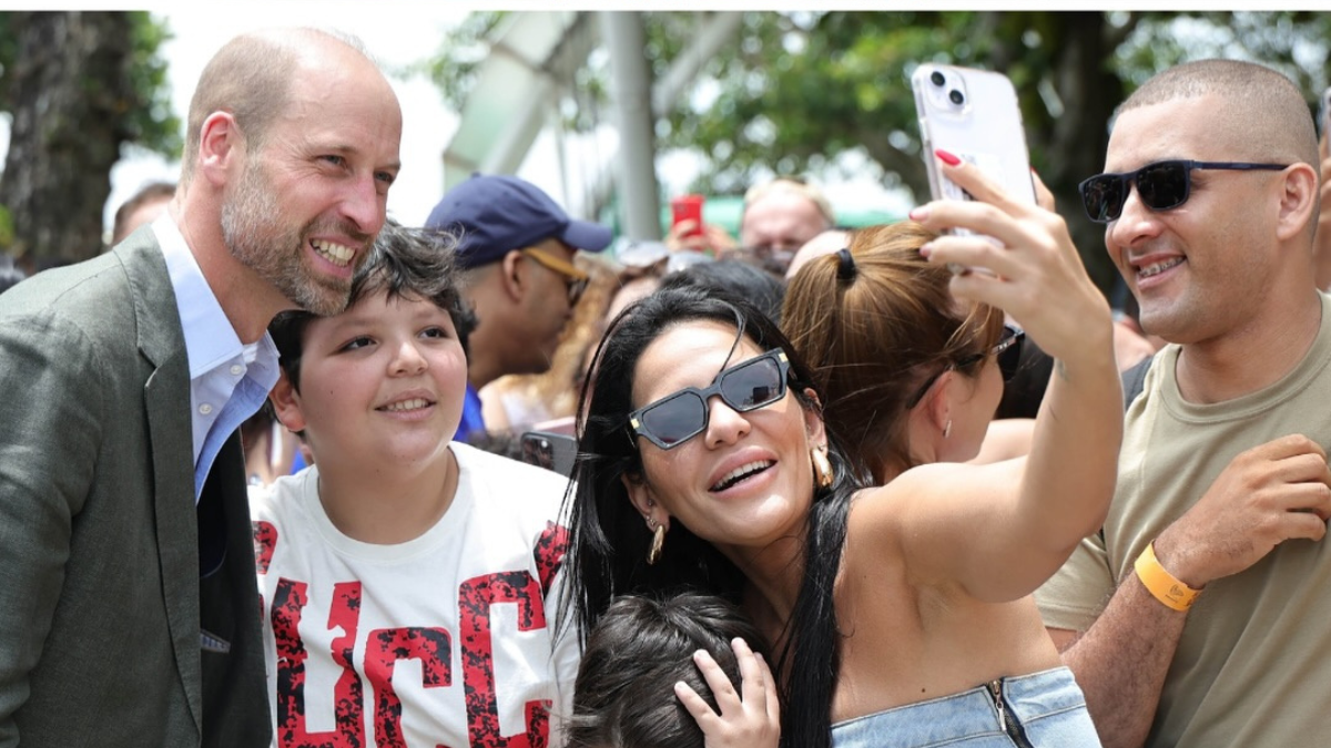 Homem sorri ao posar para uma foto com um grupo de pessoas que fazem uma selfie; uma mulher segura o celular, enquanto outros sorridentes olham para a câmera em ambiente externo e ensolarado.