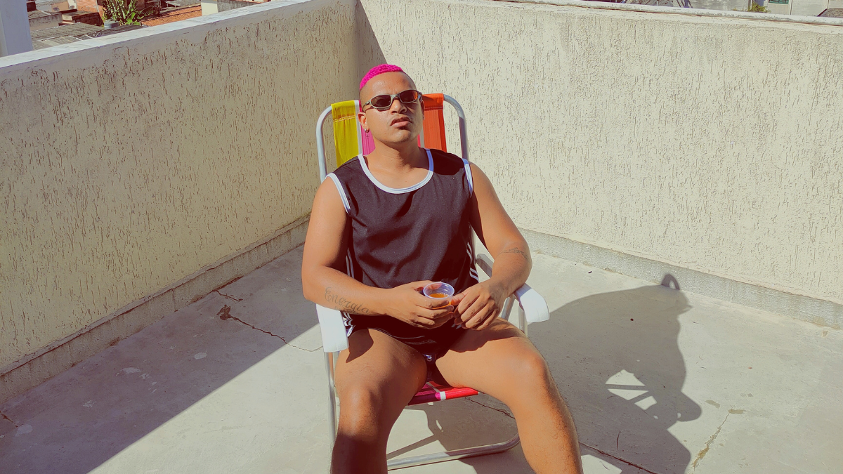 Homem com cabelo rosa e óculos escuros relaxa em uma cadeira de praia colorida, usando regata preta e segurando um copo pequeno, sob sol forte em uma área externa de piso de cimento e parede bege.