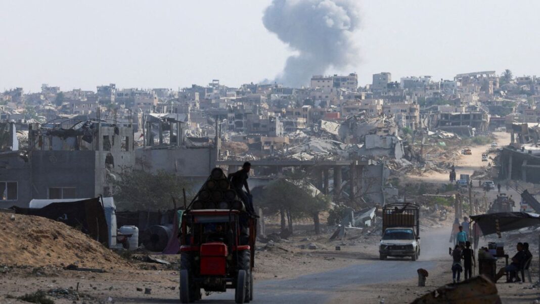 Gaza Trator carrega duas pessoas em estrada enquanto ao fundo uma fumaça grande aparece entre as casas