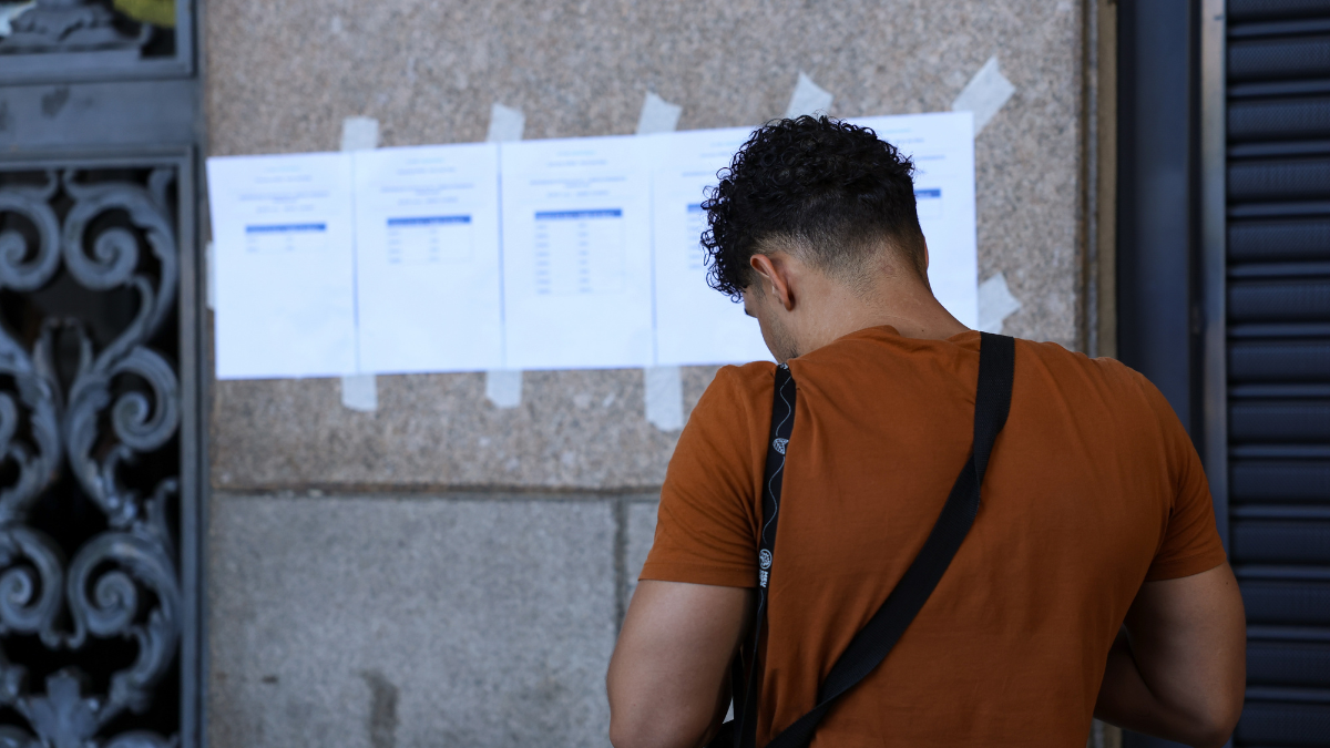 CNU_Prova_Tomaz SilvaAgência Brasil Homem de camiseta laranja e bolsa transversal preta observa folhas impressas afixadas com fita adesiva em uma parede de pedra. Os papéis contêm listas com nomes e números, possivelmente resultados ou locais de prova. A cena ocorre em ambiente urbano, em frente a um prédio público, com parte de um portão de ferro decorado visível à esquerda.