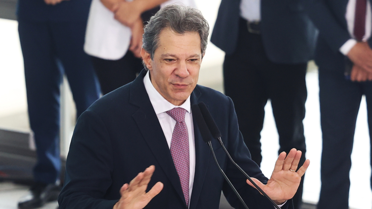 Fernando Haddad_Foto_Agência Brasil Homem de terno azul escuro, camisa branca e gravata rosa fala em um púlpito com dois microfones. Ele gesticula com as mãos levantadas à altura do peito, como se enfatizasse um ponto. Ao fundo, pessoas vestidas formalmente aparecem desfocadas, sugerindo um ambiente oficial ou de evento público.