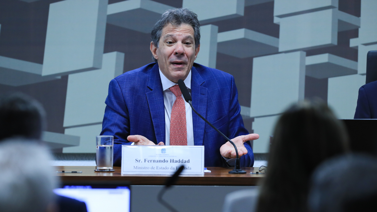 Fernando Haddad_Ministro da Fazenda_Economia_Foto_Agência Brasil Homem de terno azul e gravata vermelha fala ao microfone em uma mesa de audiência. À sua frente há uma placa com o nome “Sr. Fernando Haddad, Ministro de Estado da Fazenda”. Ele gesticula com as mãos abertas, em gesto de explicação. Ao fundo, há um painel com formas geométricas e pessoas assistindo à fala.