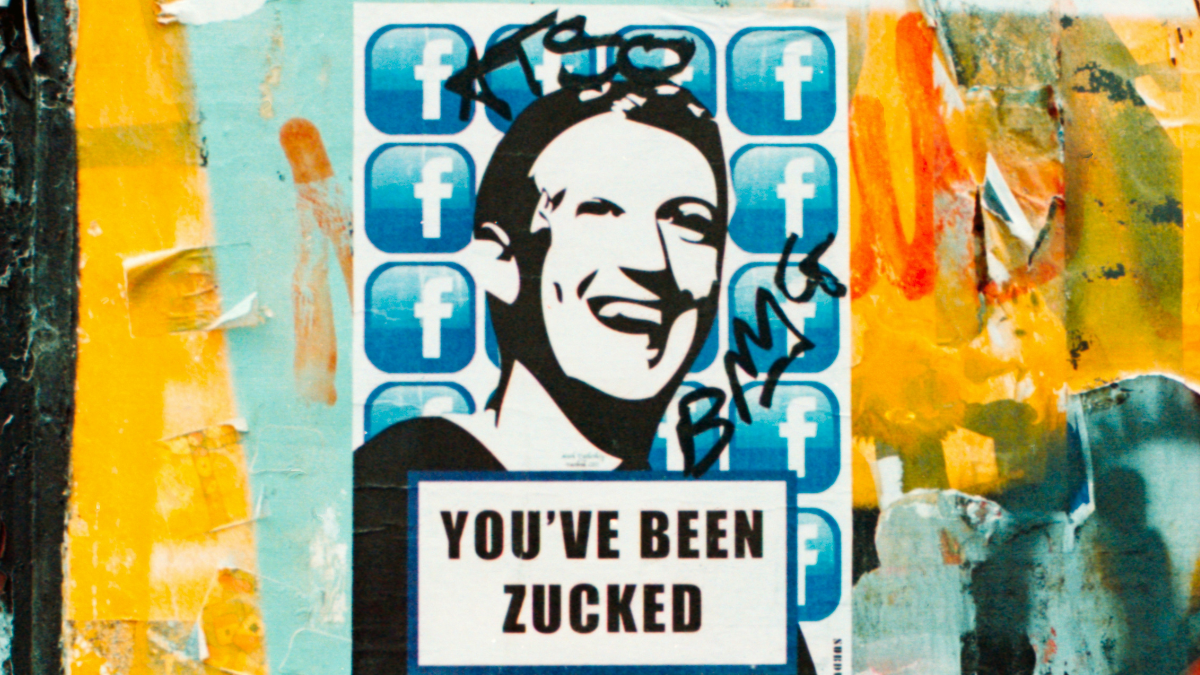 Em um parede com grafites, há um poster com uma ilustração do rosto de Mark Zuckenberg, CEO da Meta, com a seguinte frase em inglês: 
