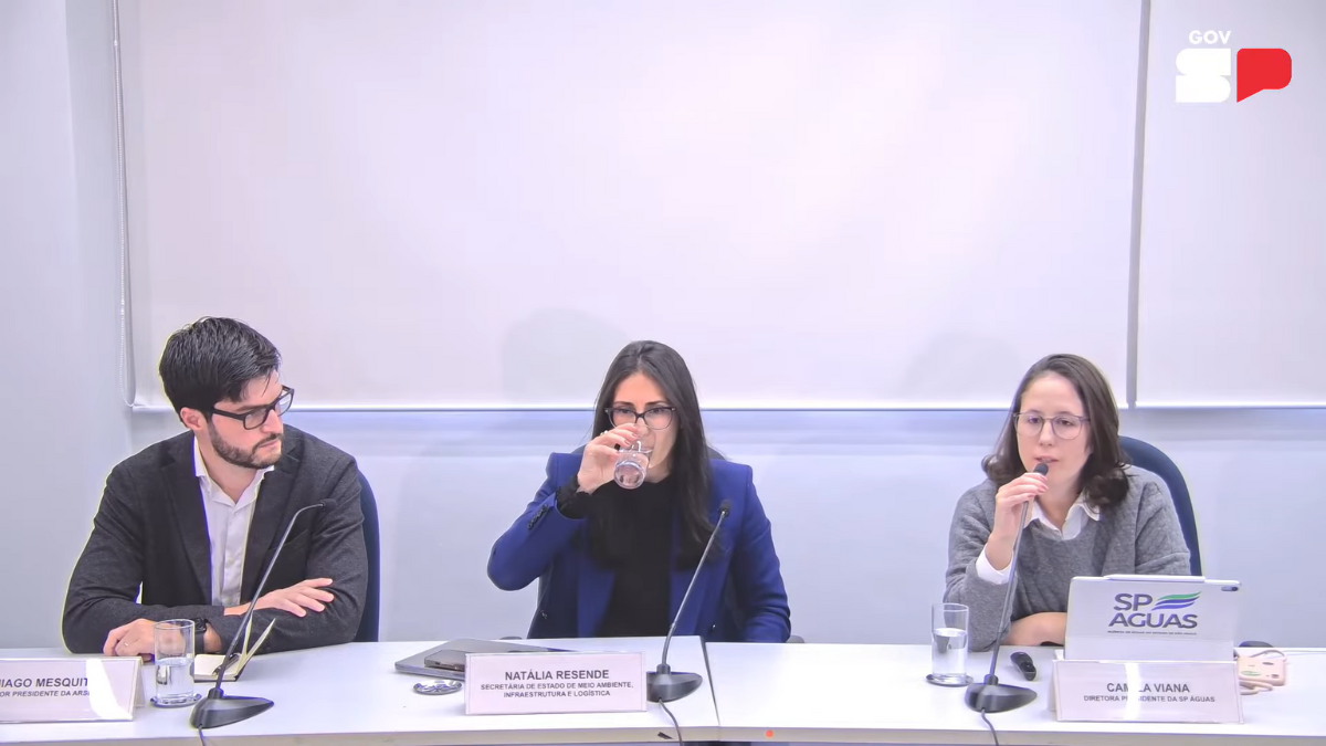 Coletiva de Impresa_Governo do Estado_Reprodução Três pessoas estão sentadas lado a lado em uma mesa de reunião, com microfones e copos de água à frente. À esquerda, um homem de barba e óculos usa terno escuro e camisa branca. No centro, uma mulher de cabelos escuros e lisos, vestindo blazer azul, bebe um copo de água. À direita, outra mulher de óculos, com suéter cinza e camisa branca, fala ao microfone. À frente dela há um notebook com o logotipo “SP Águas” e uma placa de identificação. Ao fundo, uma parede branca com o logotipo do Governo de São Paulo.