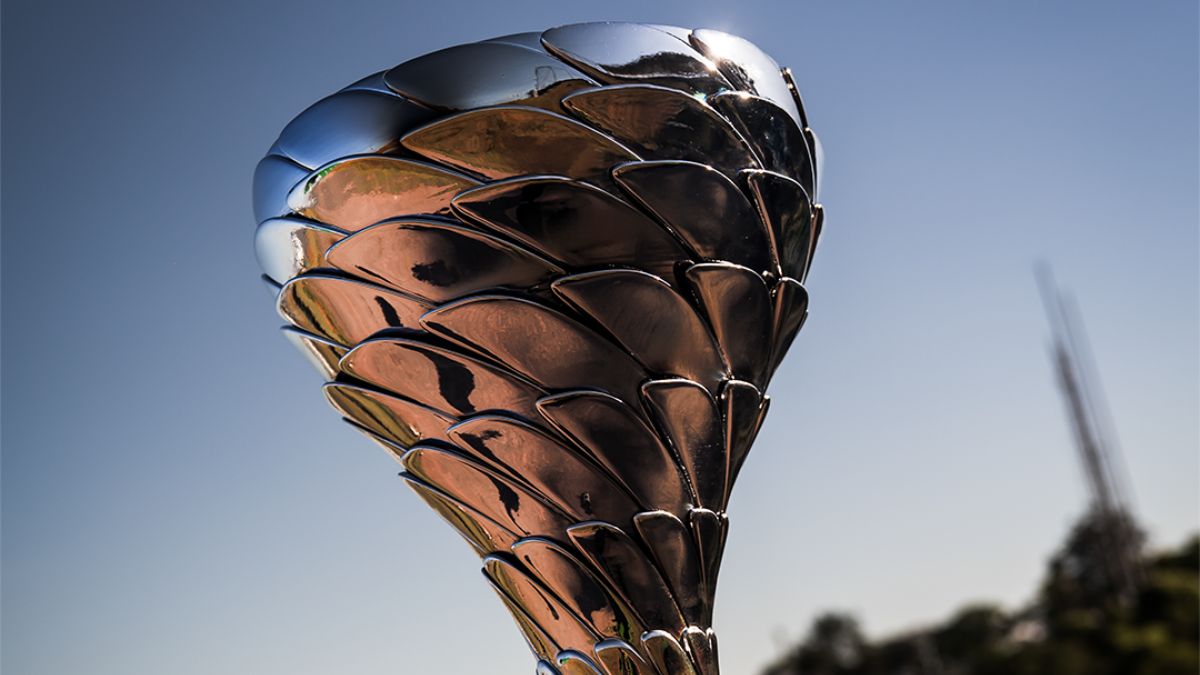 MUNDO (2) Troféu metálico prateado com design moderno, formado por placas sobrepostas que lembram escamas ou pétalas. Ele está exposto sobre uma base preta, com um autódromo ao fundo sob céu azul e luz solar intensa, sugerindo um evento de corrida ou premiação esportiva.