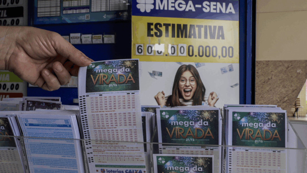 Mega da Virada 2025_Foto_Agência Brasil Pessoa segura um volante da “Mega da Virada” em uma casa lotérica. Ao fundo, um cartaz da Mega-Sena exibe a estimativa de prêmio de 600 milhões de reais e a imagem de uma mulher sorridente comemorando.