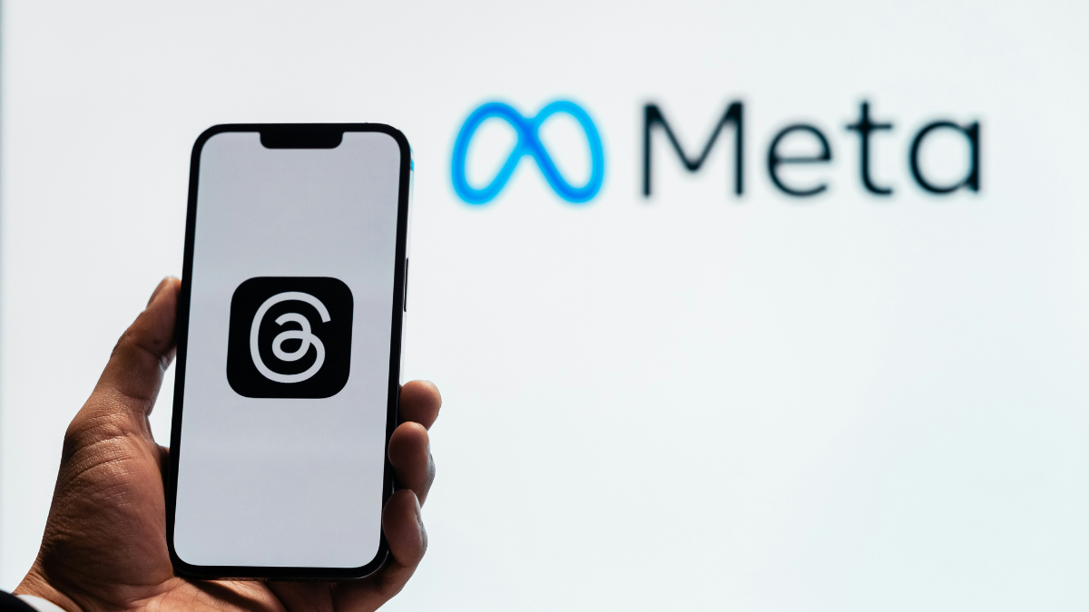 Meta_Facebook_Threads_Foto_Julio Lopez Uma pessoa segura um smartphone que exibe o logotipo do aplicativo Threads, da Meta, em fundo branco. Ao fundo, desfocado, aparece o logotipo da empresa Meta, com o símbolo azul em forma de infinito ao lado da palavra “Meta” em letras pretas. A imagem sugere o vínculo entre o aplicativo e a empresa controladora das redes sociais Facebook e Instagram.