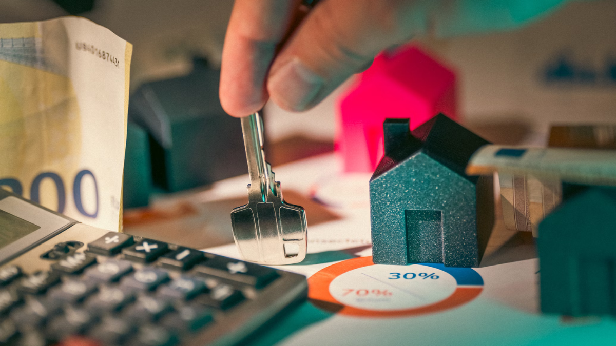 PADRÃO_IMAGEM_SITE Pessoa segura uma chave sobre uma mesa com miniaturas de casas, notas de dinheiro, calculadora e gráfico circular, representando o mercado imobiliário e o custo do aluguel.