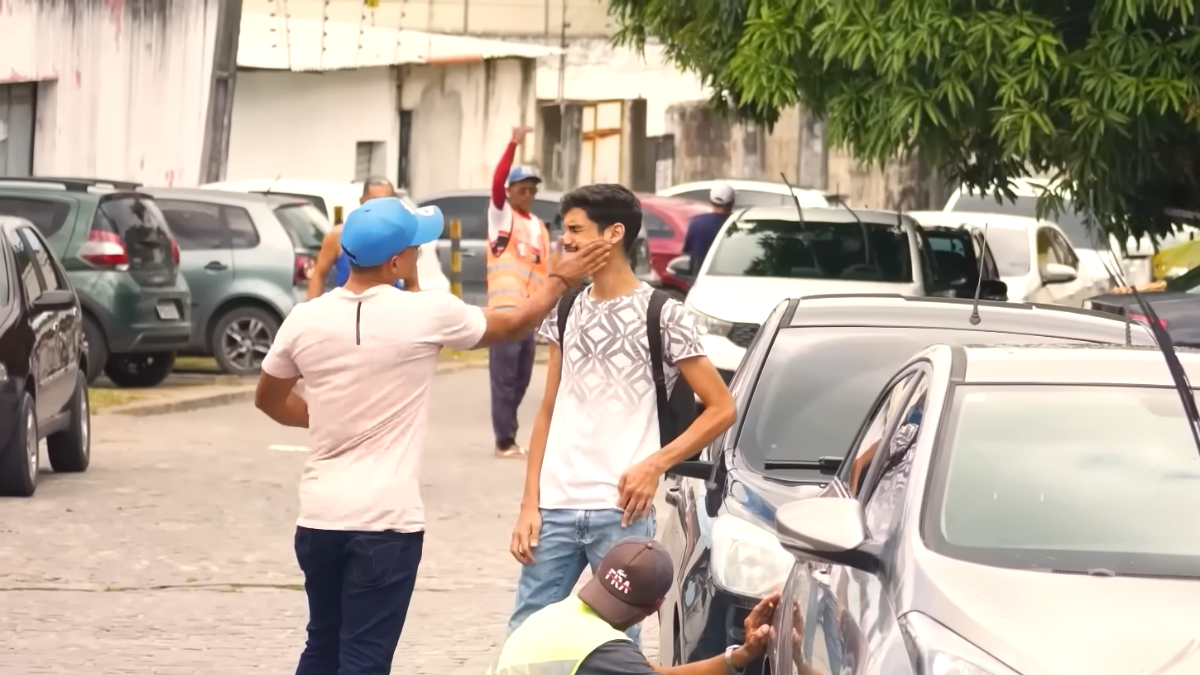Rafael Chocolate_Pegadinha_Foto_Reprodução Em uma rua com carros estacionados, um homem de boné azul e camiseta clara dá um tapa no rosto de outro homem mais jovem, que usa mochila e camiseta branca com estampa geométrica. O jovem parece surpreso e inclina o rosto para o lado. Ao fundo, há outras pessoas caminhando e um homem com colete laranja acenando com o braço levantado. A cena ocorre em ambiente urbano, durante o dia