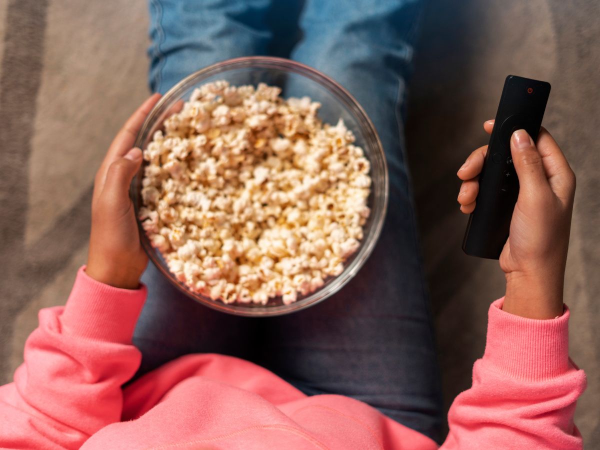 Mulher sentada no chão. Na mão ela segura um controle remoto e um pote de pipoca.