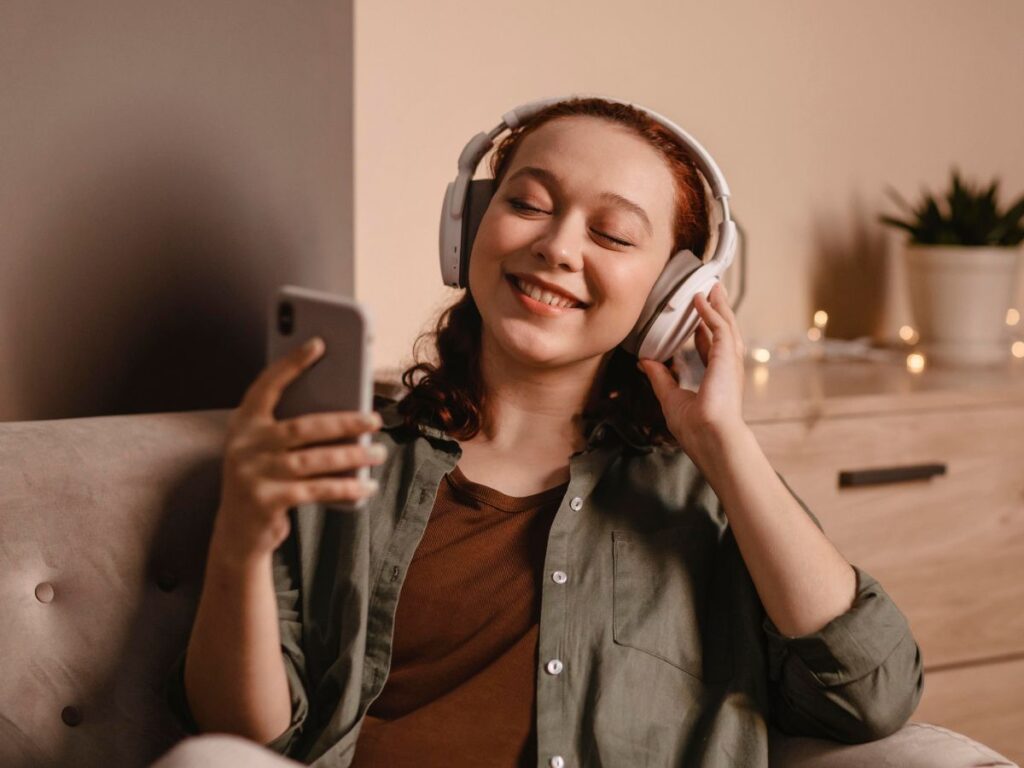 Mulher branca está com um fone de ouvido e segurando um celular em um sófa.