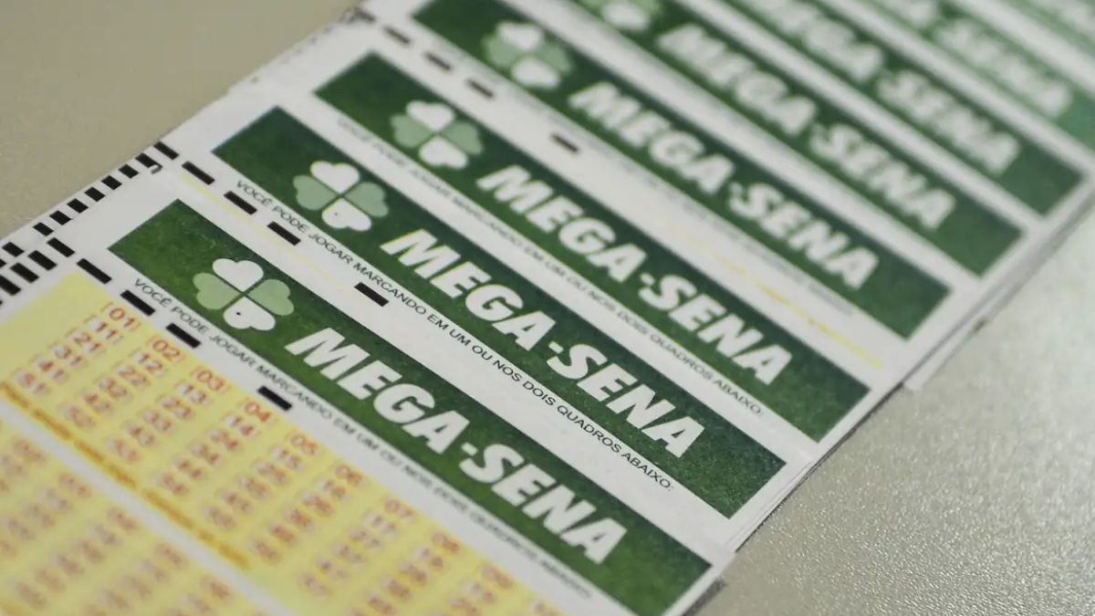 mega sena ok A imagem é um plano aproximado, em ângulo diagonal, que mostra vários bilhetes de apostas da loteria Mega-Sena. Os bilhetes estão empilhados ou dispostos lado a lado. Em primeiro plano, no canto inferior esquerdo, é possível ver as primeiras linhas da grade de números da aposta, impressas em tinta amarela e vermelha sobre o papel branco.