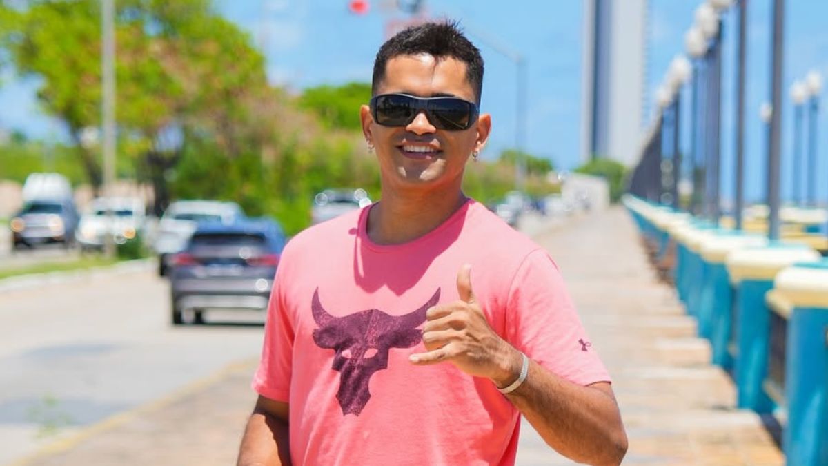 Imagem mostra o influenciador Rafael Chocolate, conhecido por fazer pegadinhas no Recife, vestido com camisa rosa, na rua, em dia ensolarado.
