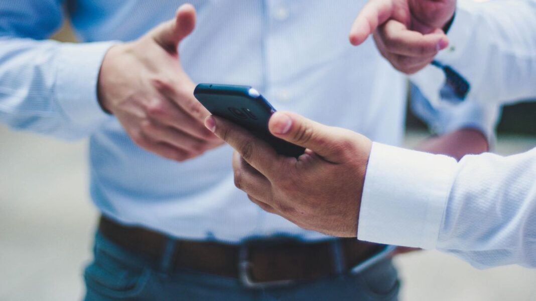 Uma mão segura um celular enquanto outra mão aponta para o aparelho