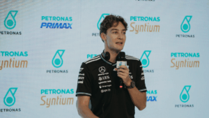 Homem veste camiseta preta da equipe Mercedes-AMG Petronas e segura um microfone durante entrevista ou coletiva de imprensa, com logotipos da Petronas e Syntium exibidos em um painel de fundo.