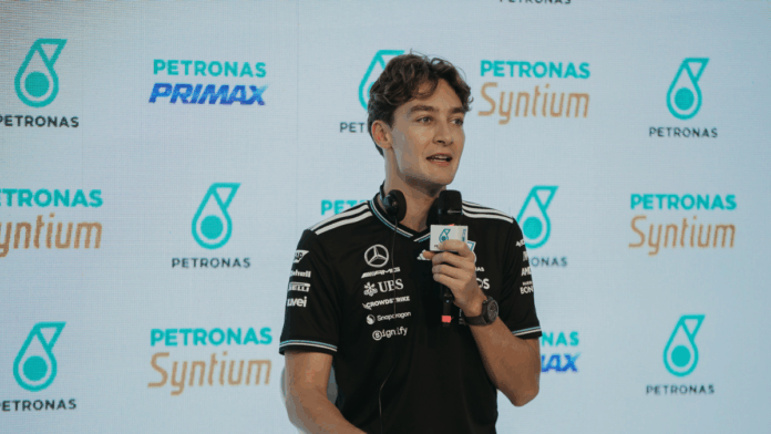 Homem veste camiseta preta da equipe Mercedes-AMG Petronas e segura um microfone durante entrevista ou coletiva de imprensa, com logotipos da Petronas e Syntium exibidos em um painel de fundo.