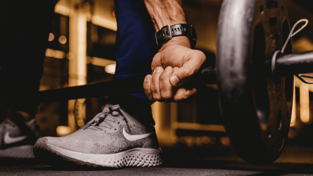 Mão masculina firme segurando uma barra de peso carregada, com foco nos músculos do antebraço e no tênis esportivo cinza, enquanto o ambiente de academia aparece desfocado ao fundo.