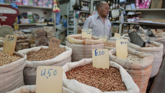 A imagem mostra uma banca de feira, com sacos de graõs e preços variados