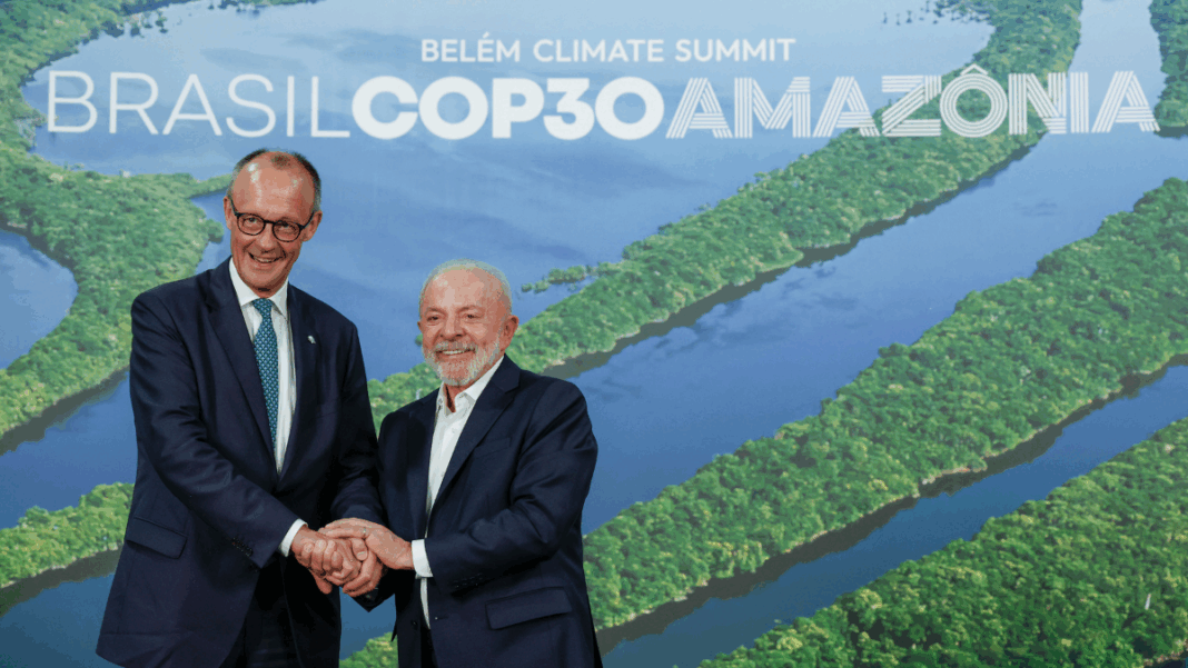 Dois homens de terno sorriem e apertam as mãos diante de um painel com imagem aérea da Floresta Amazônica e o texto “Brasil COP30 Amazônia – Belém Climate Summit”.