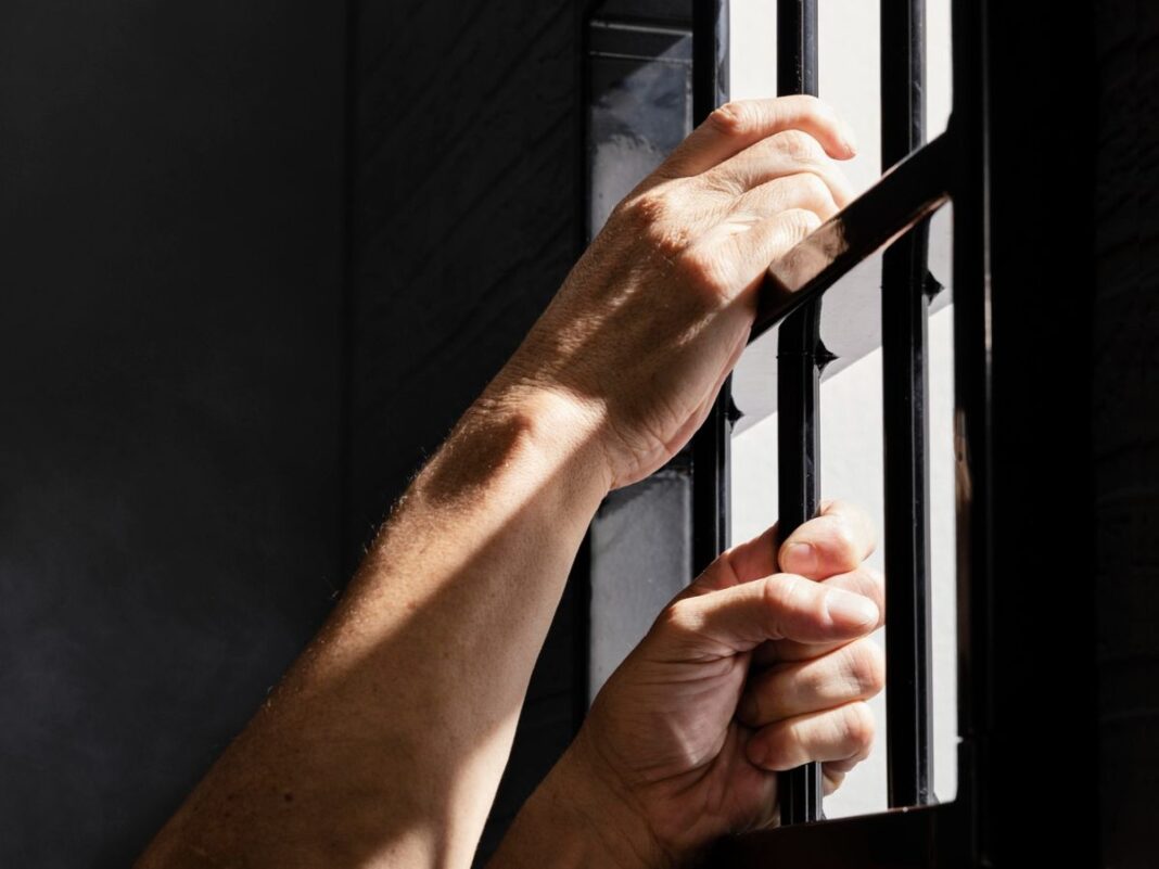 Imagem mostra as mãos de um detento segurando as grades da prisão