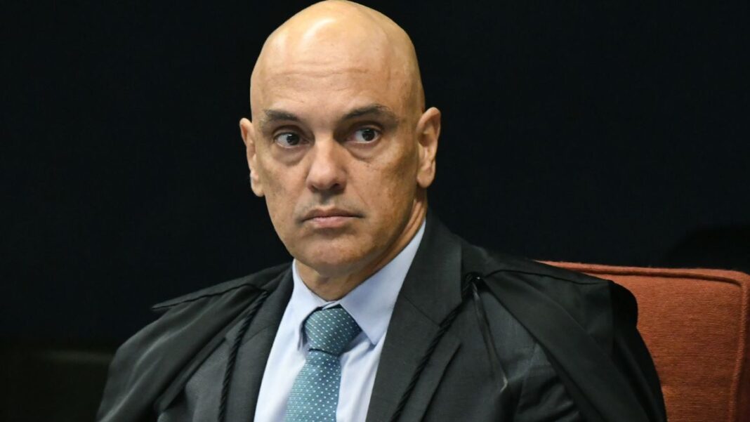 A foto é um retrato em plano médio-fechado, focado no Ministro do Supremo Tribunal Federal (STF), Alexandre de Moraes. O ministro aparece em uma pose formal, olhando diretamente para a frente ou ligeiramente para o lado direito do observador. Ele tem a cabeça raspada/calva e as sobrancelhas arqueadas, com uma expressão séria, tensa e de intensa concentração, que pode ser interpretada como desconfiança ou atenção plena. Seus olhos estão arregalados e fixos.
